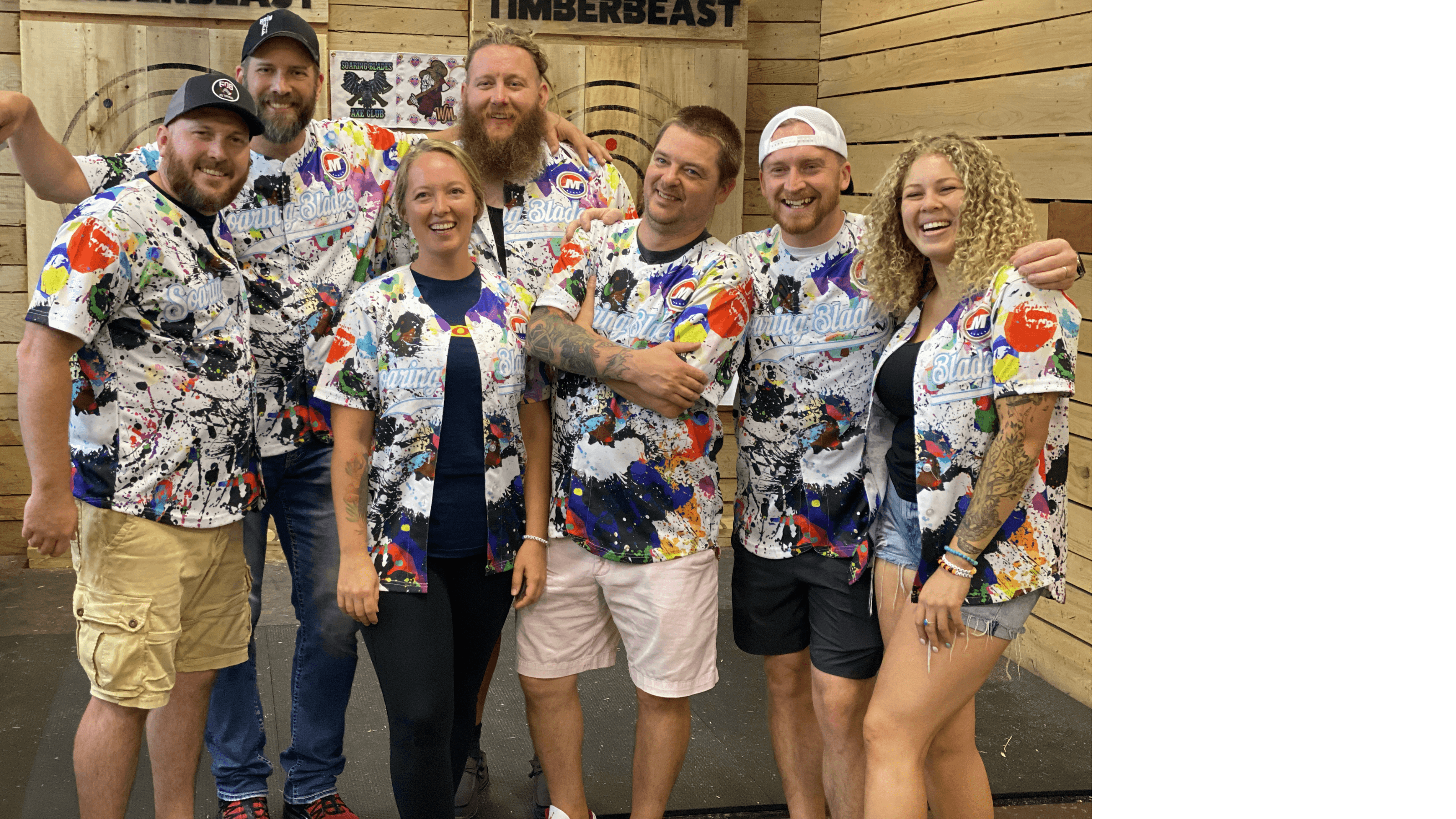 Six smiling friends in matching paint-splattered jerseys pose together at an axe-throwing venue.